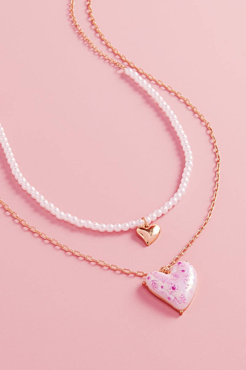 Two heart-shaped pendants on pearl necklaces against a pink background